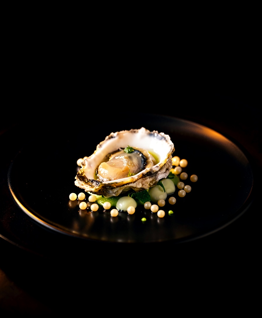 A Kumamoto oyster with green plum granité and nasturtium in a shallow bowl, dramatically lit.